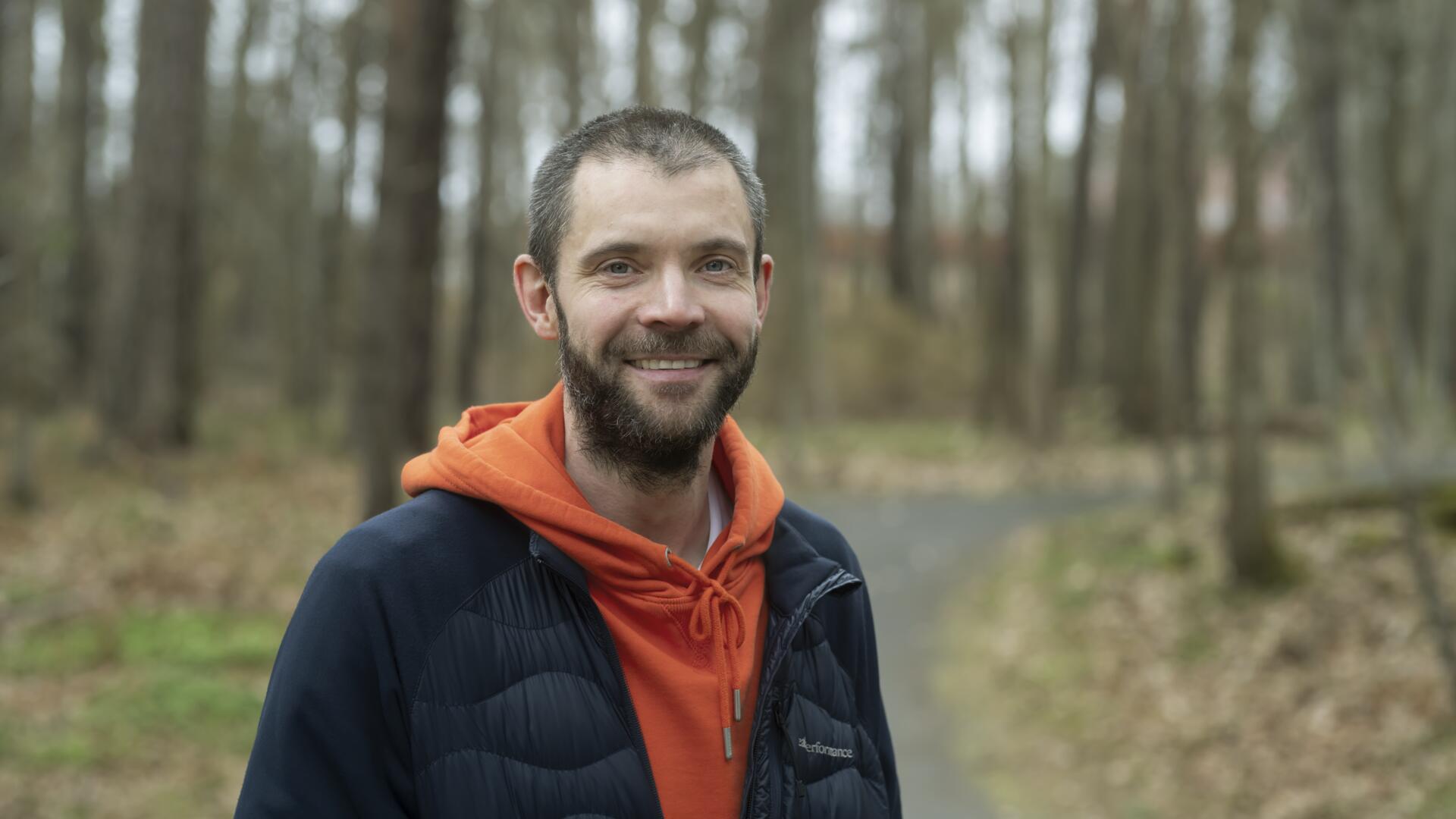 man med röd huvtröja och mörk jacka står leende längs grusväg i skogen
